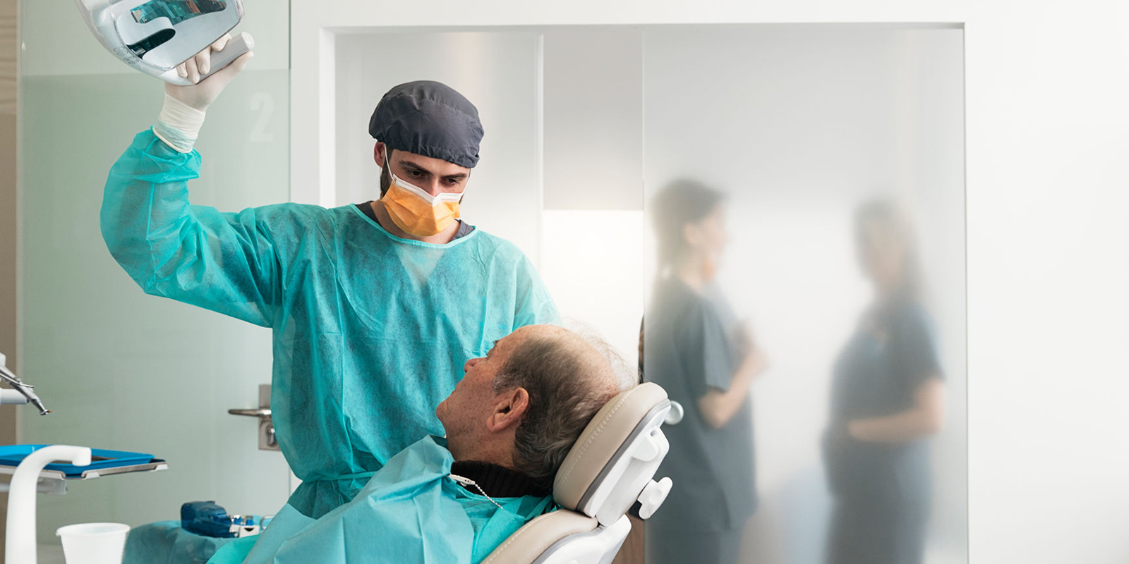 ACDQ dentiste ajustant la lampe lors d'un examen sur un patient, hygiénistes dentaires en discussion en arrière-plan